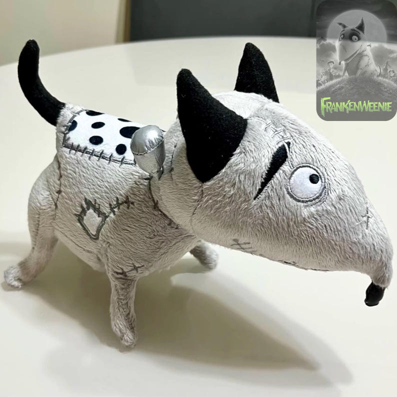 May include: A soft toy dog with a Frankenstein theme. The dog is mainly grey with black ears and tail, and a white and black spotted patch on its back. It features stitched details and a silver accessory. The image includes the text "Frankenweenie".