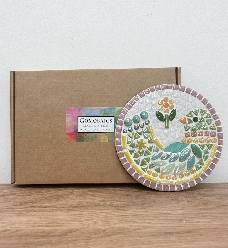 May include: A round mosaic craft kit featuring a colorful bird design with a yellow flower. The kit comes in a brown cardboard box with the company name "Gomosatics" and website address printed on a label.