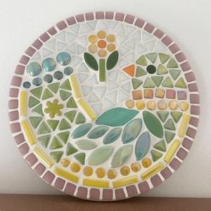 May include: A round mosaic art piece with a pink border. The mosaic features a stylized bird with green and blue leaves, a yellow flower with an orange center, and yellow and white tiles.