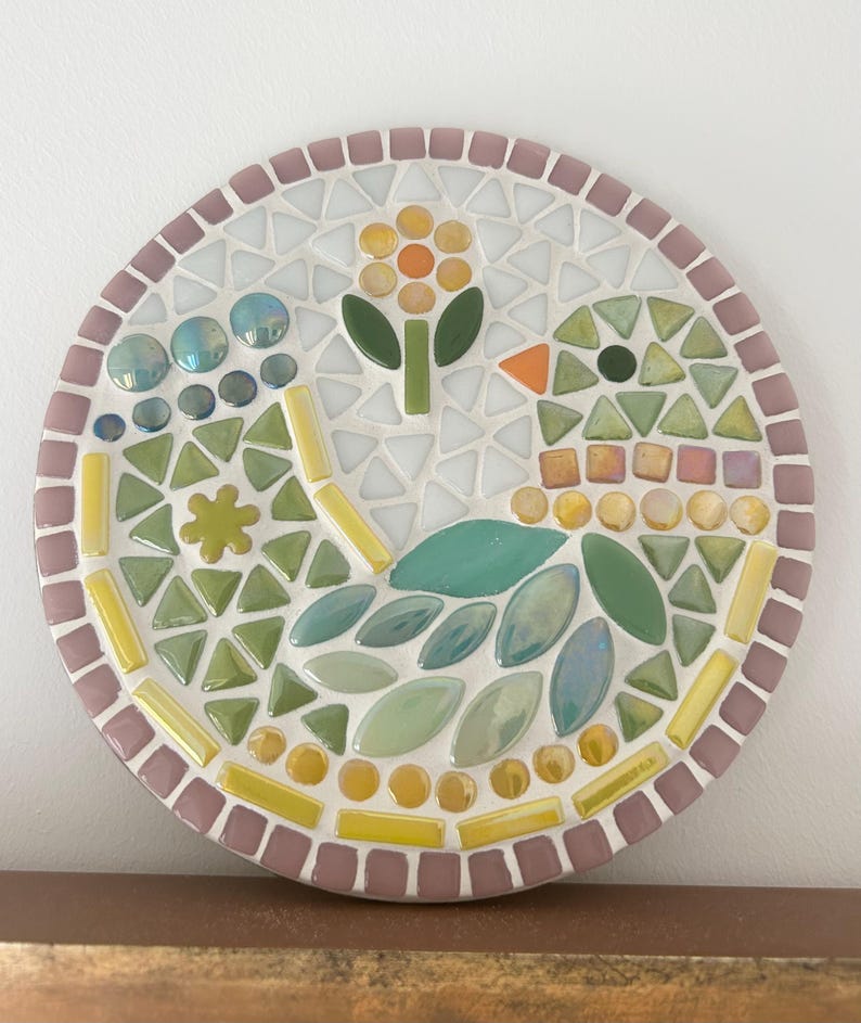 May include: A round mosaic art piece with a pink border. The mosaic features a stylized bird with green and blue leaves, a yellow flower with an orange center, and yellow and white tiles.