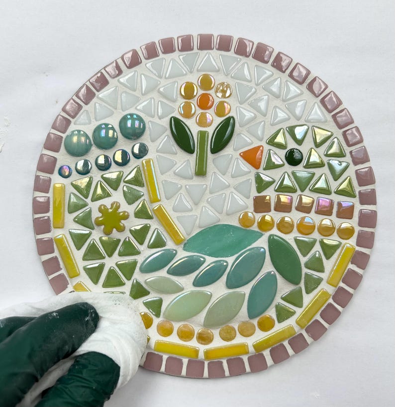May include: A round mosaic art piece with a floral design. The mosaic is made of small, colorful tiles in shades of green, yellow, orange, and white. The piece is set on a white background.