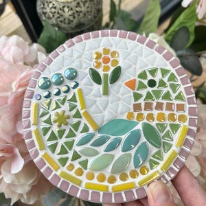 May include: A round mosaic coaster with a pink border. The design features a green leaf pattern with a yellow flower and a small bird with a green head and orange beak.