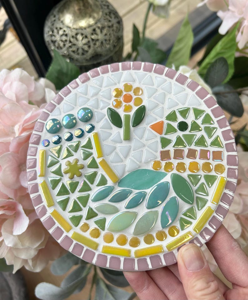 May include: A round mosaic coaster with a pink border. The design features a green leaf pattern with a yellow flower and a small bird with a green head and orange beak.