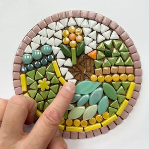 May include: A close-up of a mosaic art piece with a flower design. The flower is made of yellow, orange, and green tiles. The background is made of pink, white, and green tiles.