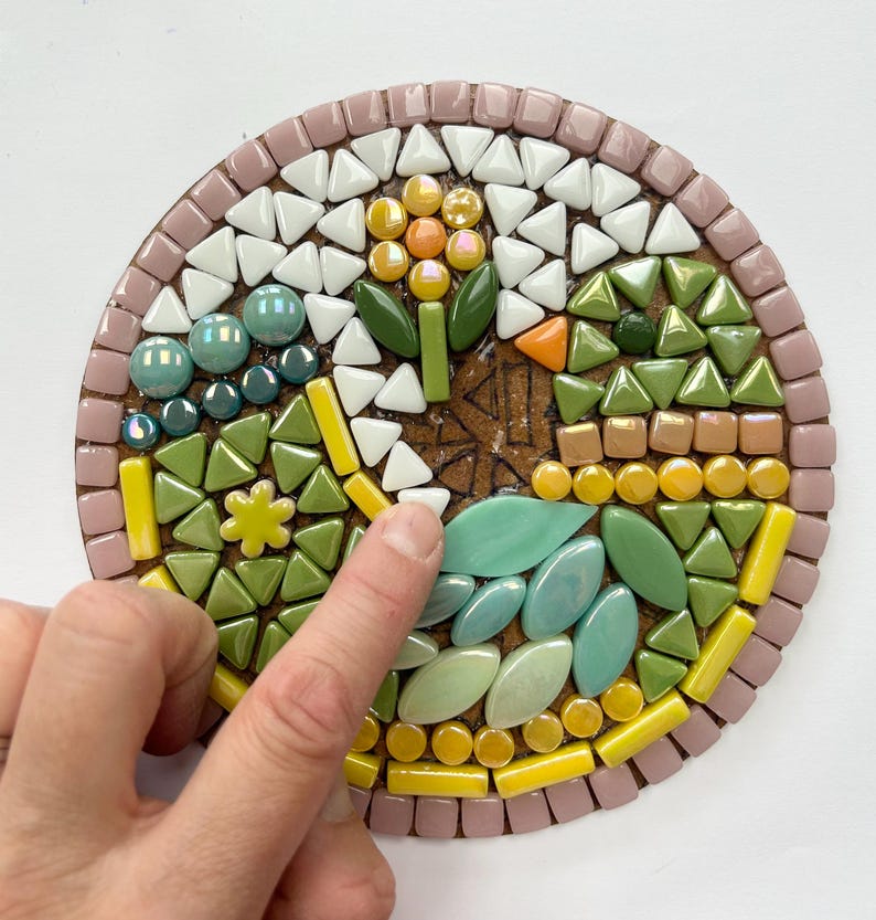 May include: A close-up of a mosaic art piece with a flower design. The flower is made of yellow, orange, and green tiles. The background is made of pink, white, and green tiles.
