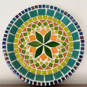 May include: A circular mosaic plate with a floral pattern. The design includes a central green flower, orange, green, and yellow geometric shapes. The outer edge is teal with iridescent square tiles.