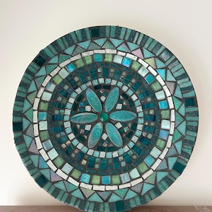 May include: A circular mosaic wall art piece featuring a central flower motif made of teal glass petals. The design is composed of various shades of blue, green, and iridescent glass tiles arranged in concentric rings with dark grout lines.