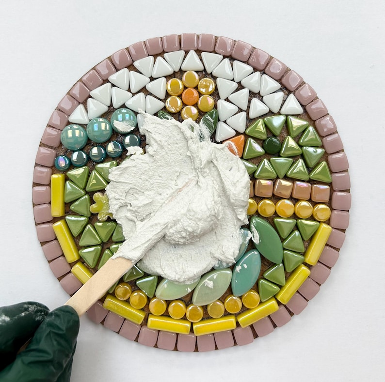 May include: A close-up of a round mosaic tile with a white, green, and yellow design. A white grout is being applied to the tile with a wooden tool.