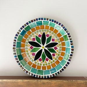 Può includere: Un piatto a mosaico circolare con un disegno centrale a fiore a sei petali in tessere di vetro verde scuro e nero. Il motivo è circondato da anelli concentrici di tessere quadrate arancioni, verde chiaro e turchesi, con un bordo iridescente.
