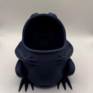 May include: A dark navy blue frog-shaped container with a wide, open mouth. The frog is depicted in a seated position with its front legs resting against its chest. The surface of the object shows a subtle, layered texture consistent with 3D printing.