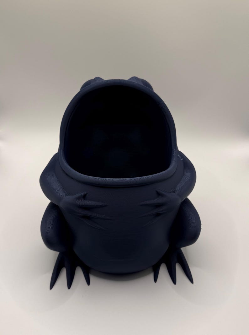 May include: A dark navy blue frog-shaped container with a wide, open mouth. The frog is depicted in a seated position with its front legs resting against its chest. The surface of the object shows a subtle, layered texture consistent with 3D printing.