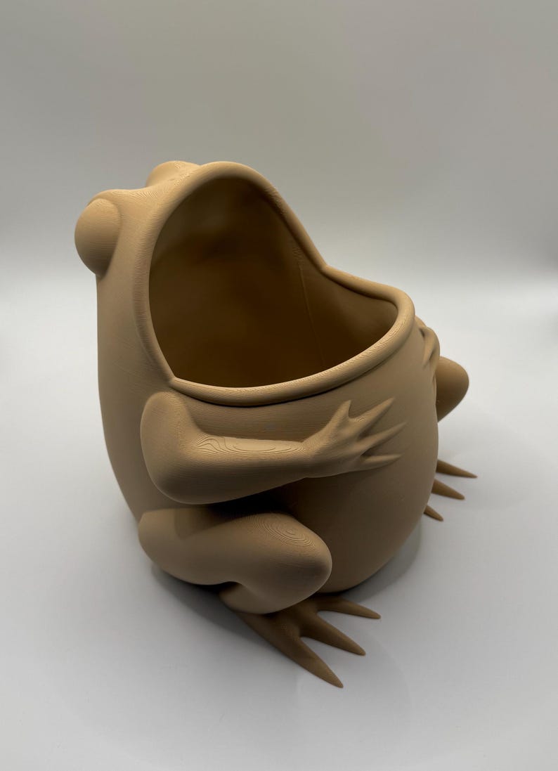 May include: A light tan, matte-finished frog-shaped container with a wide, open top. The object features a smooth, rounded body with sculpted limbs, webbed feet, and prominent eyes, showing a visible 3D-printed texture across its surface.