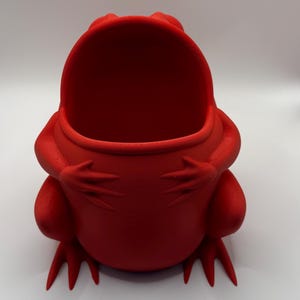 May include: A bright red, hollow frog-shaped container with a wide, open mouth. The frog is depicted in a seated position with its front limbs resting against its rounded belly. The surface of the object shows a fine, layered texture throughout.