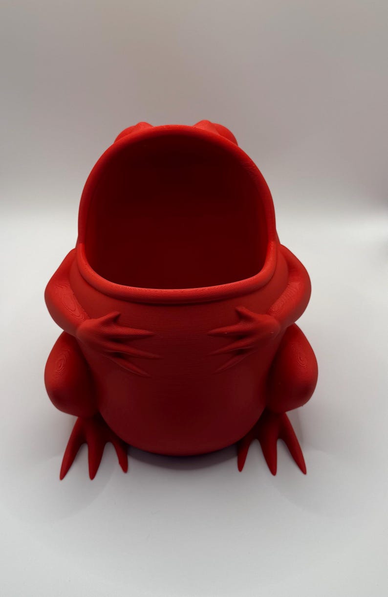 May include: A bright red, hollow frog-shaped container with a wide, open mouth. The frog is depicted in a seated position with its front limbs resting against its rounded belly. The surface of the object shows a fine, layered texture throughout.