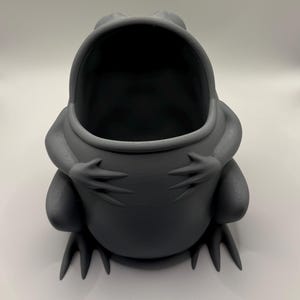 May include: A matte dark gray, hollow frog-shaped container with a wide, open mouth. The frog is depicted in a seated position with its front limbs resting on its belly, featuring visible 3D-printed layer lines across its smooth, rounded surface.