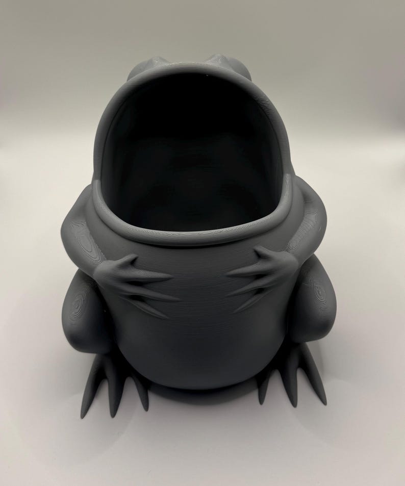 May include: A matte dark gray, hollow frog-shaped container with a wide, open mouth. The frog is depicted in a seated position with its front limbs resting on its belly, featuring visible 3D-printed layer lines across its smooth, rounded surface.