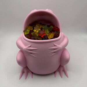 May include: A light pink, frog-shaped container with a wide, open mouth. The interior of the frog is filled with a collection of multi-colored, star-shaped gummy candies in shades of red, yellow, orange, and green against a plain background.