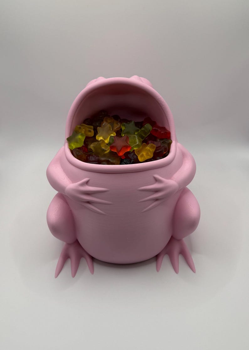 May include: A light pink, frog-shaped container with a wide, open mouth. The interior of the frog is filled with a collection of multi-colored, star-shaped gummy candies in shades of red, yellow, orange, and green against a plain background.