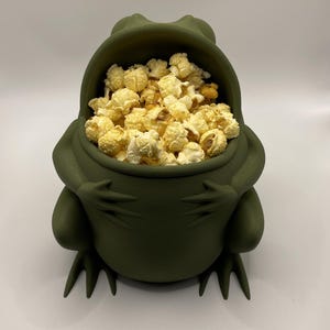 May include: A matte olive green bowl shaped like a seated frog with its arms crossed over its belly. The open mouth of the frog serves as the bowl's opening, which is filled with a generous portion of yellow, buttery popcorn kernels.
