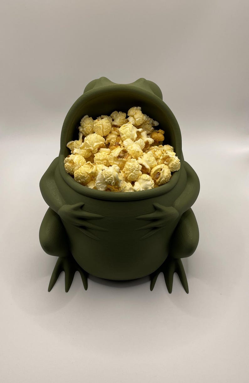May include: A matte olive green bowl shaped like a seated frog with its arms crossed over its belly. The open mouth of the frog serves as the bowl's opening, which is filled with a generous portion of yellow, buttery popcorn kernels.
