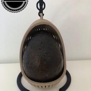 Könnte beinhalten: Eine braune Avocado in einem beigen Avocado-Halter mit einem schwarzen Metallhaken und Ständer.