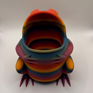 May include: A hollow, frog-shaped container featuring a vibrant, multi-colored gradient pattern with horizontal stripes of orange, yellow, blue, and purple. The frog has a wide, open mouth, visible front limbs resting on its body, and pointed feet.