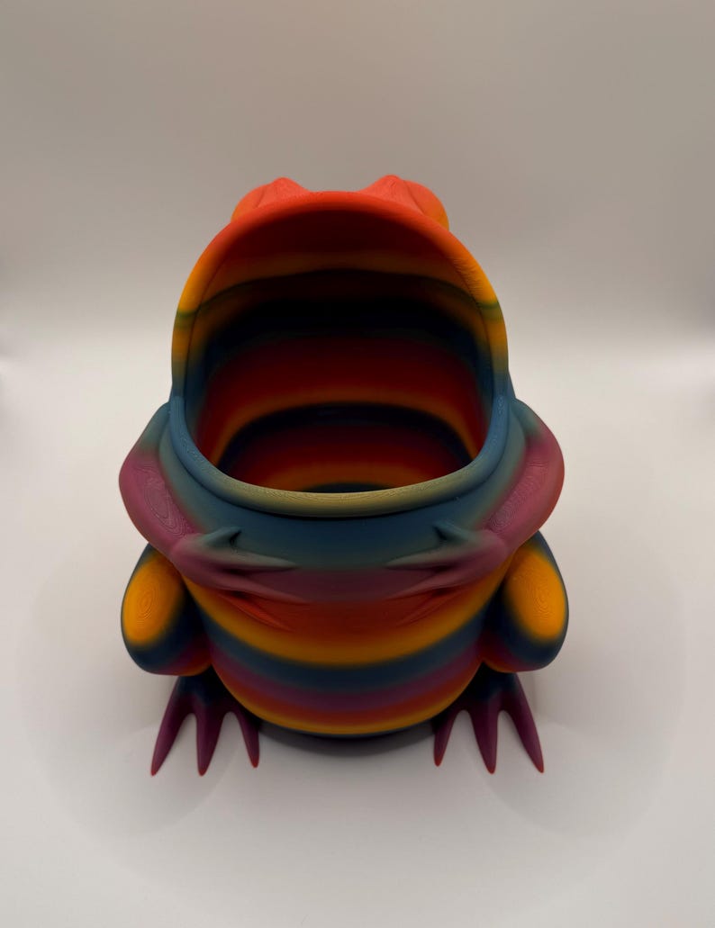 May include: A hollow, frog-shaped container featuring a vibrant, multi-colored gradient pattern with horizontal stripes of orange, yellow, blue, and purple. The frog has a wide, open mouth, visible front limbs resting on its body, and pointed feet.