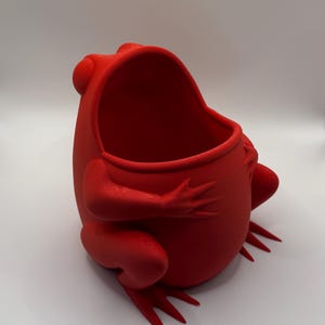 May include: A bright red, hollow frog-shaped container with a wide, open mouth. The object features a smooth, matte finish and is sculpted in a seated position with visible front and back legs extending outward from the rounded, central body cavity.