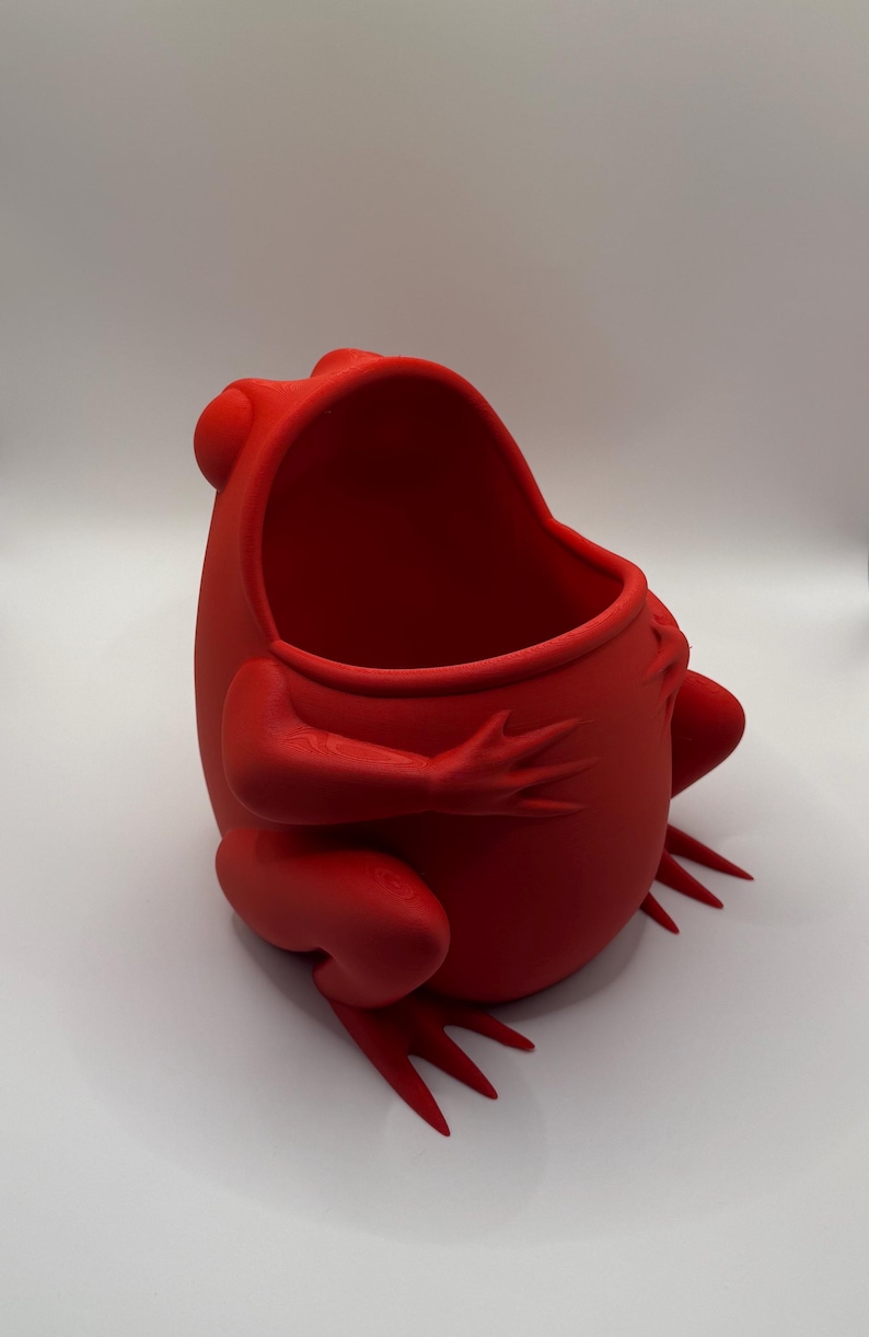 May include: A bright red, hollow frog-shaped container with a wide, open mouth. The object features a smooth, matte finish and is sculpted in a seated position with visible front and back legs extending outward from the rounded, central body cavity.