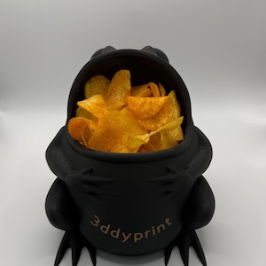 May include: A matte black, frog-shaped bowl with a wide, open mouth filled with golden-orange potato chips. The front of the bowl features the text 3ddyprint in a light brown font. The frog has visible limbs and webbed feet at the base.