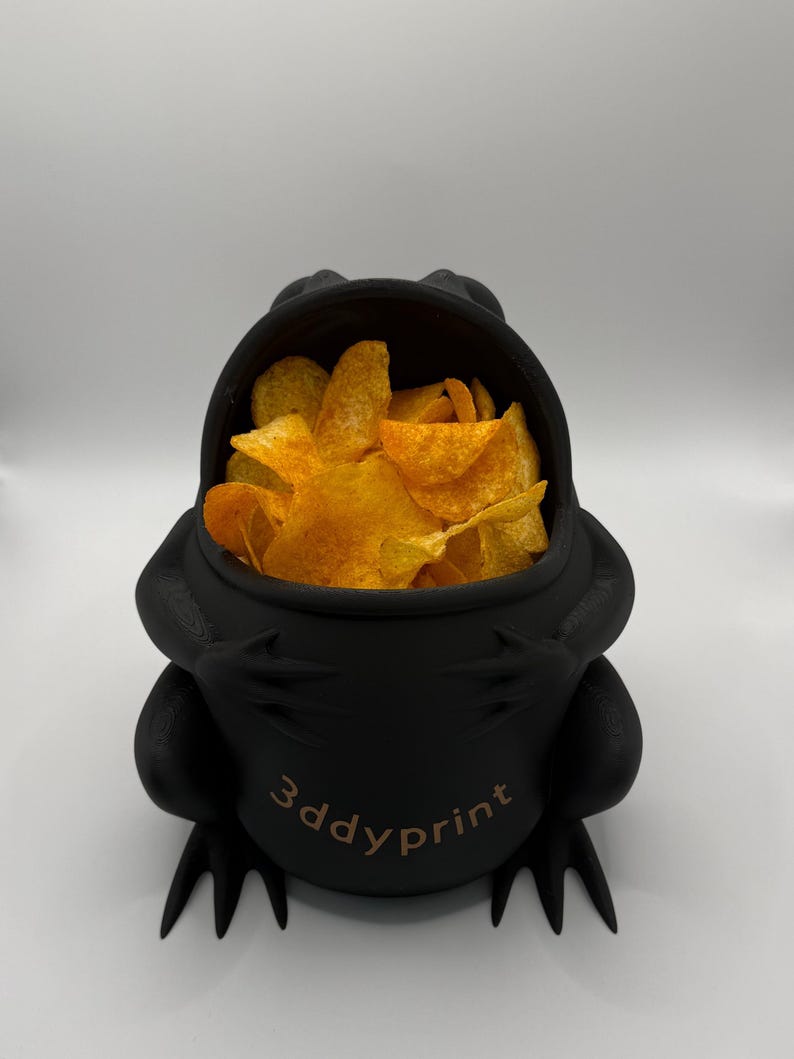 May include: A matte black, frog-shaped bowl with a wide, open mouth filled with golden-orange potato chips. The front of the bowl features the text 3ddyprint in a light brown font. The frog has visible limbs and webbed feet at the base.