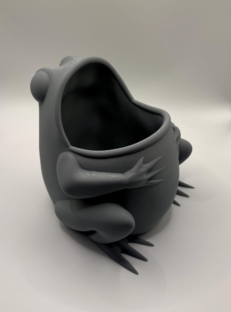 May include: A matte dark gray 3D printed container shaped like a sitting frog with a wide, open mouth. The frog has prominent eyes, defined limbs, and webbed feet, with a hollow interior space accessible through the top opening of the head.