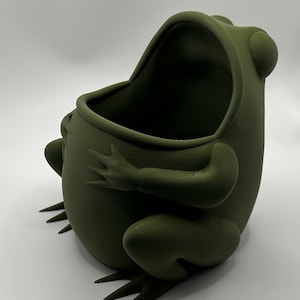 May include: A matte olive green frog-shaped container with a wide, open mouth. The frog is depicted in a seated position with its arms resting on its rounded belly and its webbed feet extending forward, featuring a smooth, uniform surface texture.