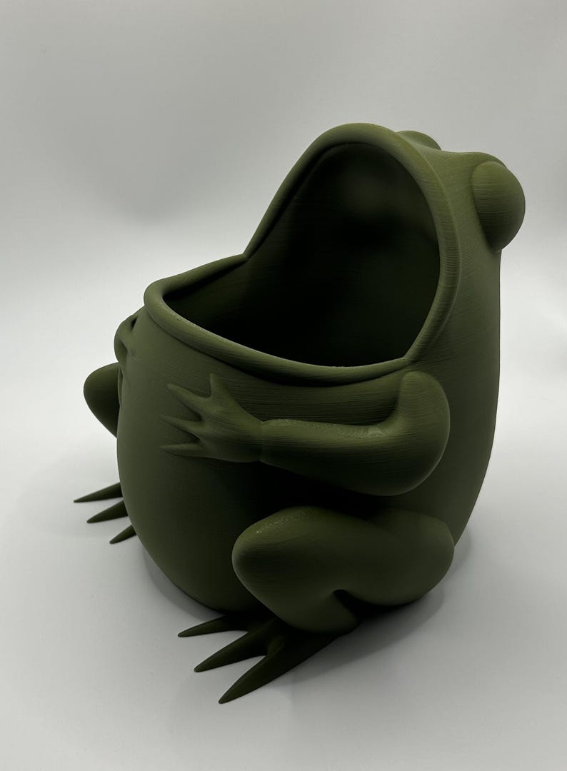 May include: A matte olive green frog-shaped container with a wide, open mouth. The frog is depicted in a seated position with its arms resting on its rounded belly and its webbed feet extending forward, featuring a smooth, uniform surface texture.