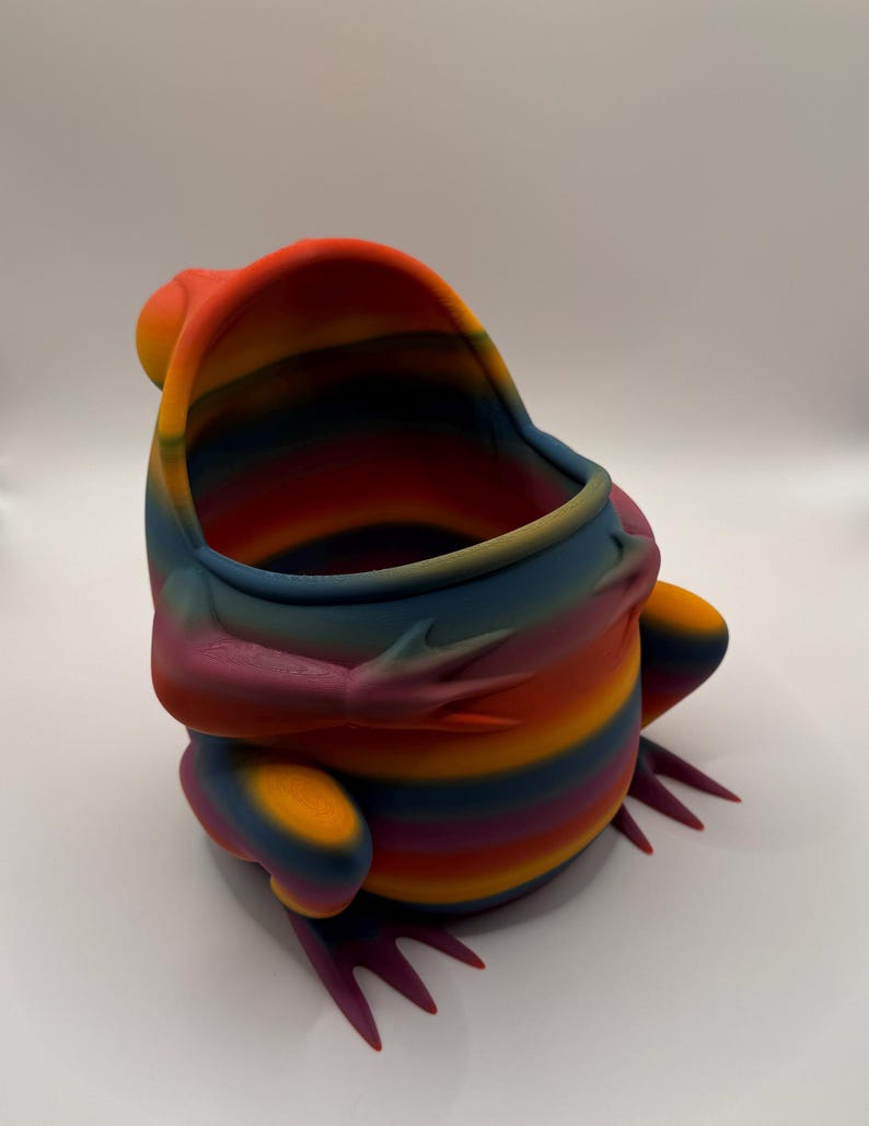 May include: A hollow, frog-shaped container featuring a vibrant, multi-colored gradient pattern with horizontal stripes in shades of orange, red, purple, blue, and yellow. The object has a wide, open top, defined limbs, and webbed feet.