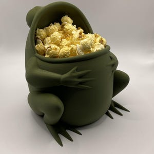 Könnte beinhalten: Ein grüner froschförmiger Behälter gefüllt mit Popcorn. Der Behälter hat einen weit geöffneten Mund und ausgestreckte Arme, wobei das Popcorn herausquillt. Der Frosch hat detaillierte Füße und eine strukturierte Oberfläche.