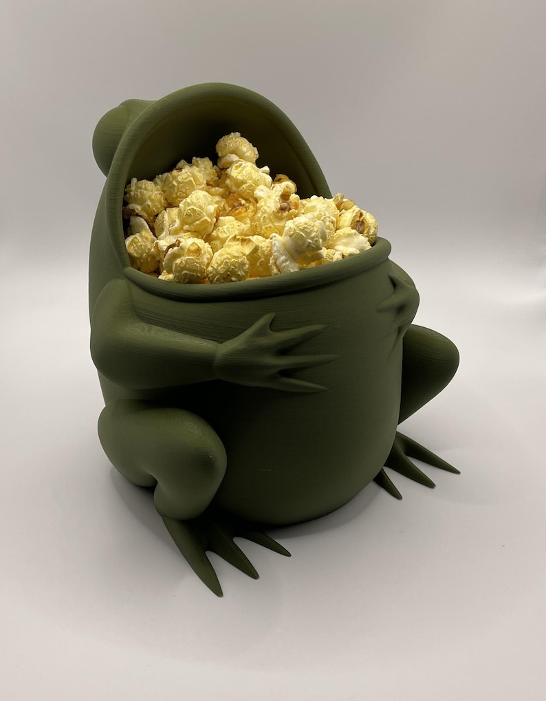 May include: A matte olive green frog-shaped bowl with a wide, open mouth serving as the container opening. The bowl is filled with a generous portion of light yellow, popped popcorn kernels, showcasing the frog's limbs and rounded body shape.