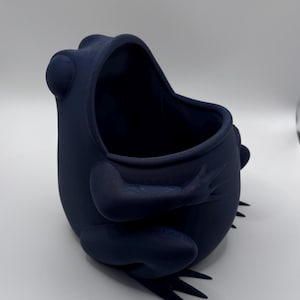 May include: A dark navy blue frog-shaped container with a smooth, matte finish. The vessel features a wide, open top, rounded body, and sculpted limbs that wrap around the sides. The frog has prominent eyes and webbed feet at the base.