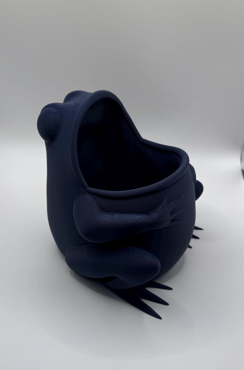 May include: A dark navy blue frog-shaped container with a smooth, matte finish. The vessel features a wide, open top, rounded body, and sculpted limbs that wrap around the sides. The frog has prominent eyes and webbed feet at the base.
