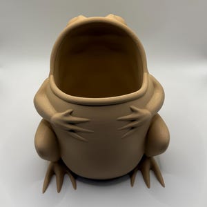 May include: A light tan, matte-finished frog-shaped container with a wide, open mouth. The frog is depicted in a seated position with its front limbs resting against its rounded belly, featuring visible 3D-printed layer lines across its surface.