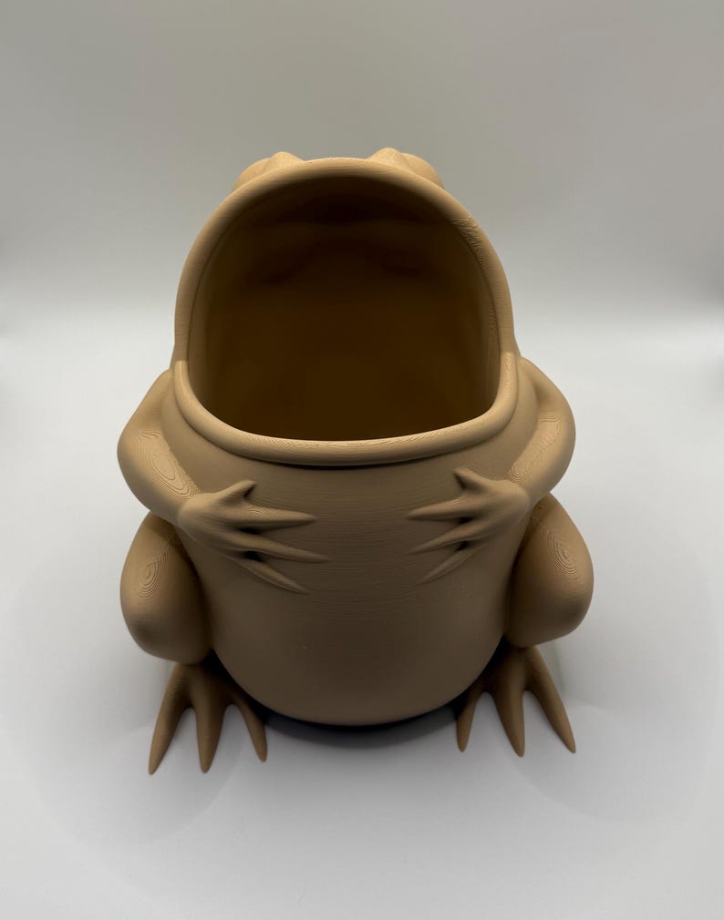 May include: A light tan, matte-finished frog-shaped container with a wide, open mouth. The frog is depicted in a seated position with its front limbs resting against its rounded belly, featuring visible 3D-printed layer lines across its surface.