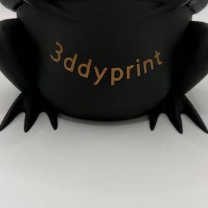 May include: A matte black, three-dimensional frog-shaped object with webbed feet and a rounded body. The text 3ddyprint is printed in a light brown color across the center of the frog's torso, featuring a smooth, solid surface texture.