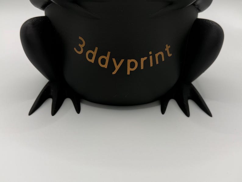 May include: A matte black, three-dimensional frog-shaped object with webbed feet and a rounded body. The text 3ddyprint is printed in a light brown color across the center of the frog's torso, featuring a smooth, solid surface texture.