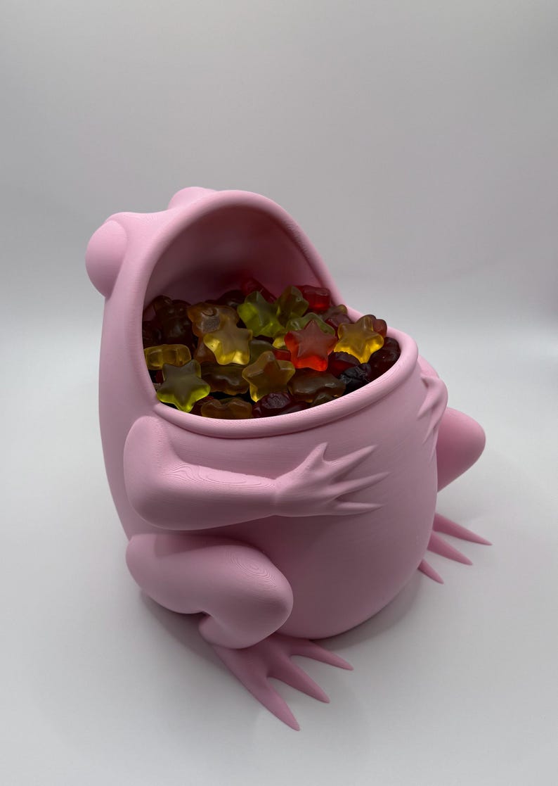 May include: A matte light pink frog-shaped container with a wide, open mouth serving as a bowl. The interior of the frog is filled with a collection of small, translucent, star-shaped gummy candies in various colors including red, yellow, and dark purple.