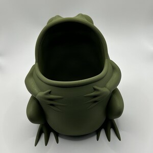May include: A matte olive green frog-shaped container with a wide, open mouth. The frog is depicted in a seated position with its front limbs resting on its belly and its hind feet extending forward, creating a hollow interior space for storage.