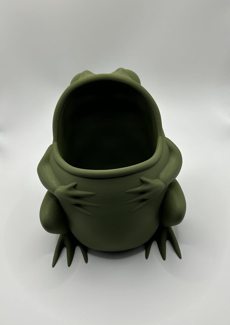 May include: A matte olive green frog-shaped container with a wide, open mouth. The frog is depicted in a seated position with its front limbs resting on its belly and its hind feet extending forward, creating a hollow interior space for storage.