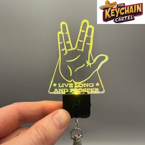 Op de afbeelding: Een heldere acryl sleutelhanger met het Vulcan-handgebaar, verlicht met een gele gloed. De tekst "LIVE LONG AND PROSPER" staat onder de hand. De sleutelhanger heeft een zwarte basis en een zilveren sleutelring.