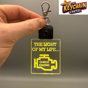 Może przedstawiać: Żółty brelok LED z tekstem "The Light of My Life..." i stylizowanym ikoną kontrolki silnika z tekstem "Check Engine".