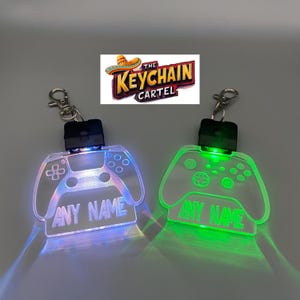 May include: Two clear acrylic video game controller keychains with light up features. The keychain on the left is lit up in blue and white, and the keychain on the right is lit up in green. Both keychains have the text "ANY NAME" engraved on them.