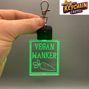 May include: A square, glowing green keychain with the words "VEGAN WANKER" and a carrot illustration. The keychain has a silver clip and a black base. The Keychain Cartel logo is in the upper right corner.